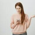 Upset or irritated ginger girl gesturing while looking at smartphone screen, being clueless or